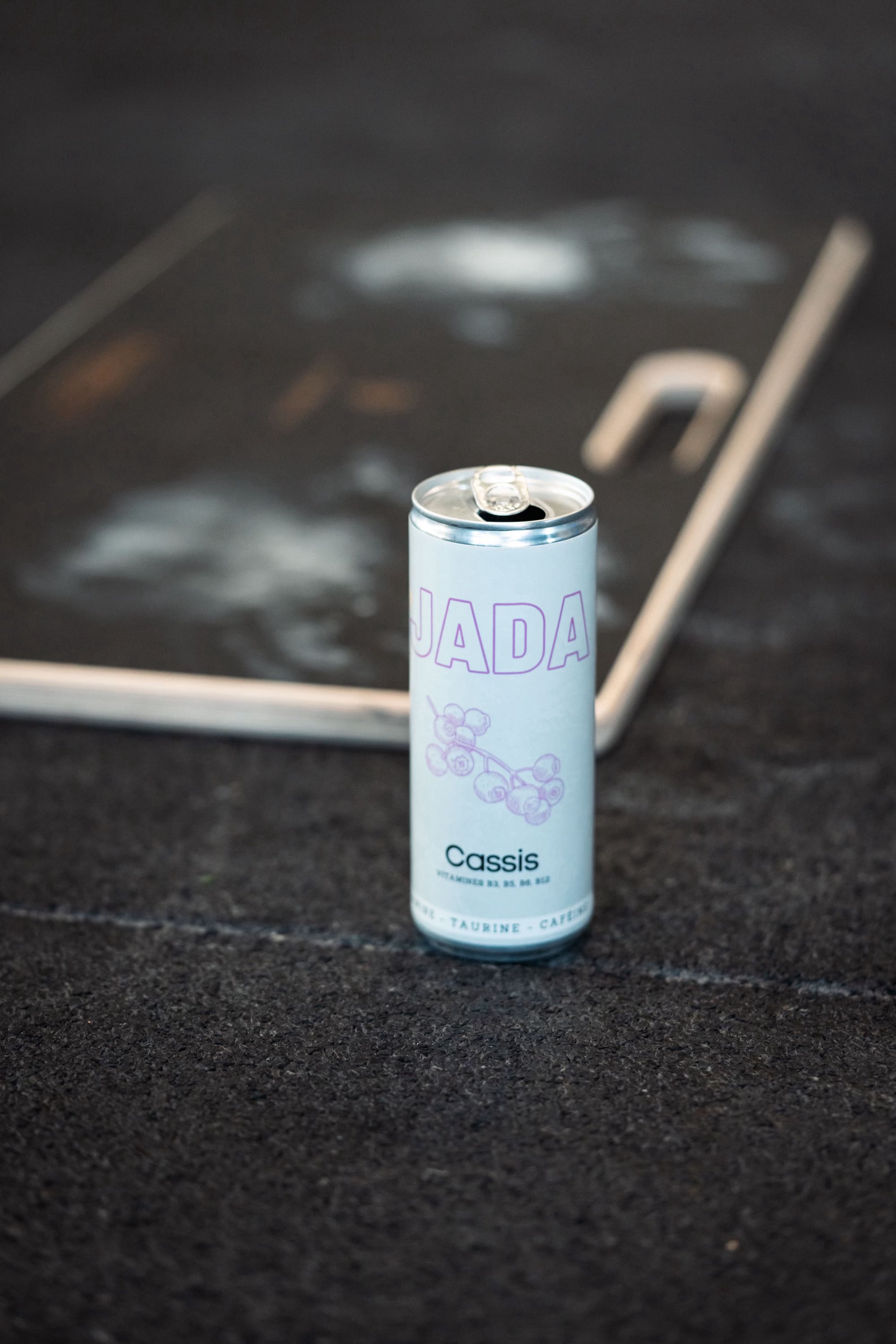 Canette ouverte de boisson énergisante JADA au cassis posée sur un trottoir urbain, avec des vitamines B3, B5, B6, B12, taurine et caféine pour un boost d'énergie instantané.